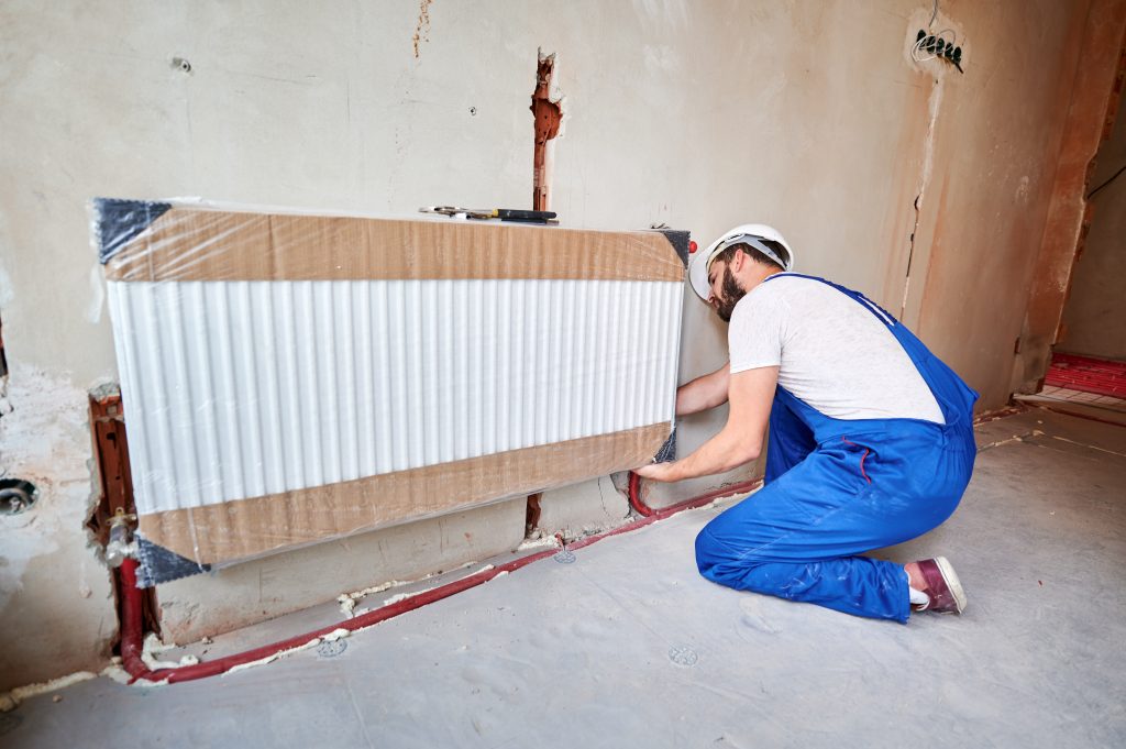 Bearded male technician in blue overalls installing a radiator in an unfinished commercial space, emphasizing HVAC heating repair services in Brooklyn.