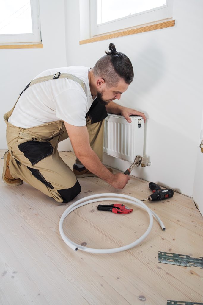 Plumber installing a new heating radiator in a commercial space, with tools and piping on the floor, emphasizing HVAC maintenance services.