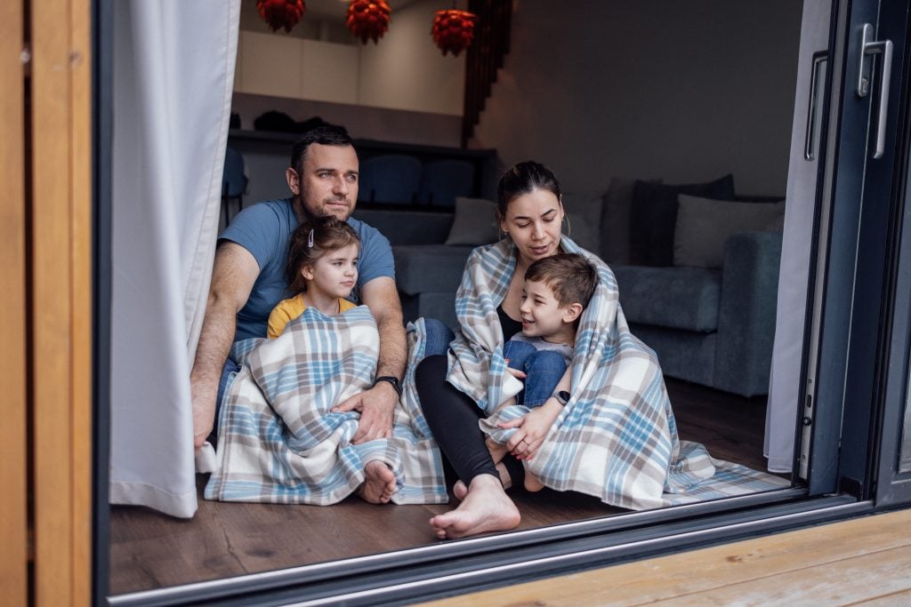 Family of four sitting on the floor, wrapped in blankets, enjoying time together indoors, with a cozy living room setting in the background, reflecting comfort and warmth.